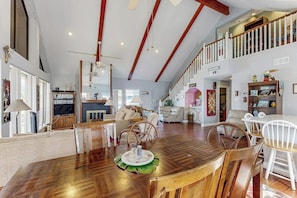 Uma sala de jantar ampla com mesa e cadeiras de madeira, teto alto com vigas aparentes e vista para a sala de estar.