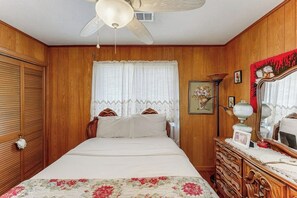 Quarto com cama, paredes de madeira, ventoinha no teto, janela com cortinas de renda, cômoda com espelho e um quadro na parede.