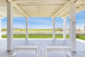 A covered outdoor area with a wooden deck, white benches, and a view of a grassy field and fence. - Blue Galveston Getaway (Galveston)