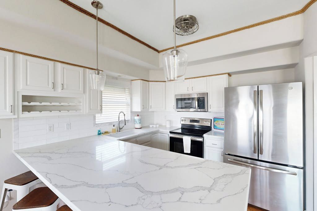 Una cocina moderna con armarios blancos, una isla de mármol, electrodomésticos de acero inoxidable e iluminación colgante.