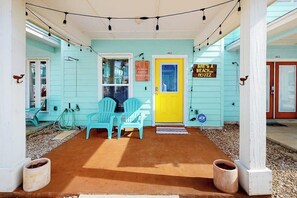 A porch with two turquoise chairs, a yellow door, and a sign that reads 'She’s a Beach... Hoozz.' - Mapacebo - Pool & Walk to Beach (Port Aransas)