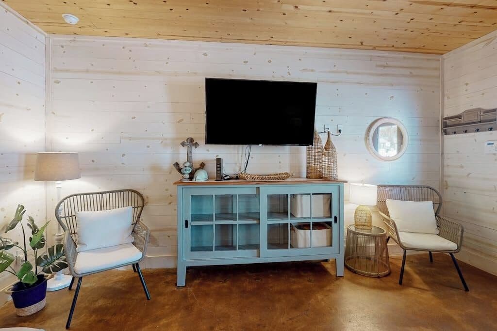 A room with a wooden ceiling, a mounted flat-screen TV, a light blue cabinet with glass doors, two wicker chairs with white cushions, and a round mirror on the wall.