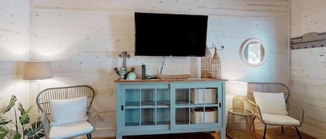 A room with a wooden ceiling, a mounted flat-screen TV, a light blue cabinet with glass doors, two wicker chairs with white cushions, and a round mirror on the wall.