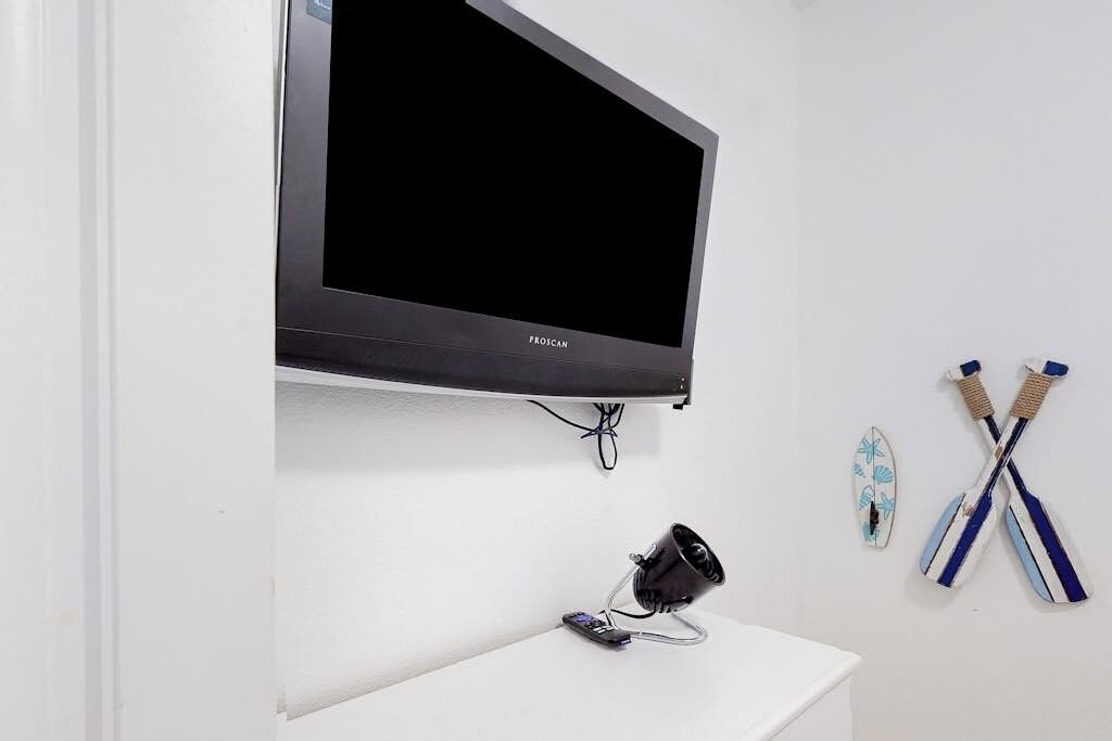 Ein an einer weißen Wand befestigter Fernseher, ein Headset auf einem Ständer und zwei Stehpaddles, die an der Wand lehnen.