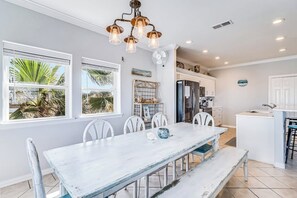 A dining area with a white table and chairs, a chandelier, and a view of palm trees through large windows. - Gulf Breezes Entire Home (Port Aransas)