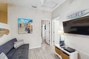 A living room with a grey sofa, a painting of surfers, a 'SURF' sign, and a flat-screen TV. - Tiki House - Ocean Views, Pool, Steps to Beach (Corpus Christi)