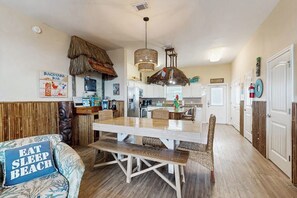 Un comedor con mesa y sillas de madera, una lámpara colgante y una cocina al fondo.