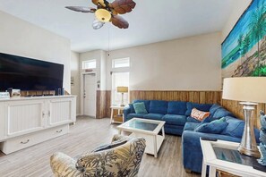 A living room with a television, a blue sofa, a coffee table, and a ceiling fan. - Tiki House - Ocean Views, Pool, Steps to Beach (Corpus Christi)