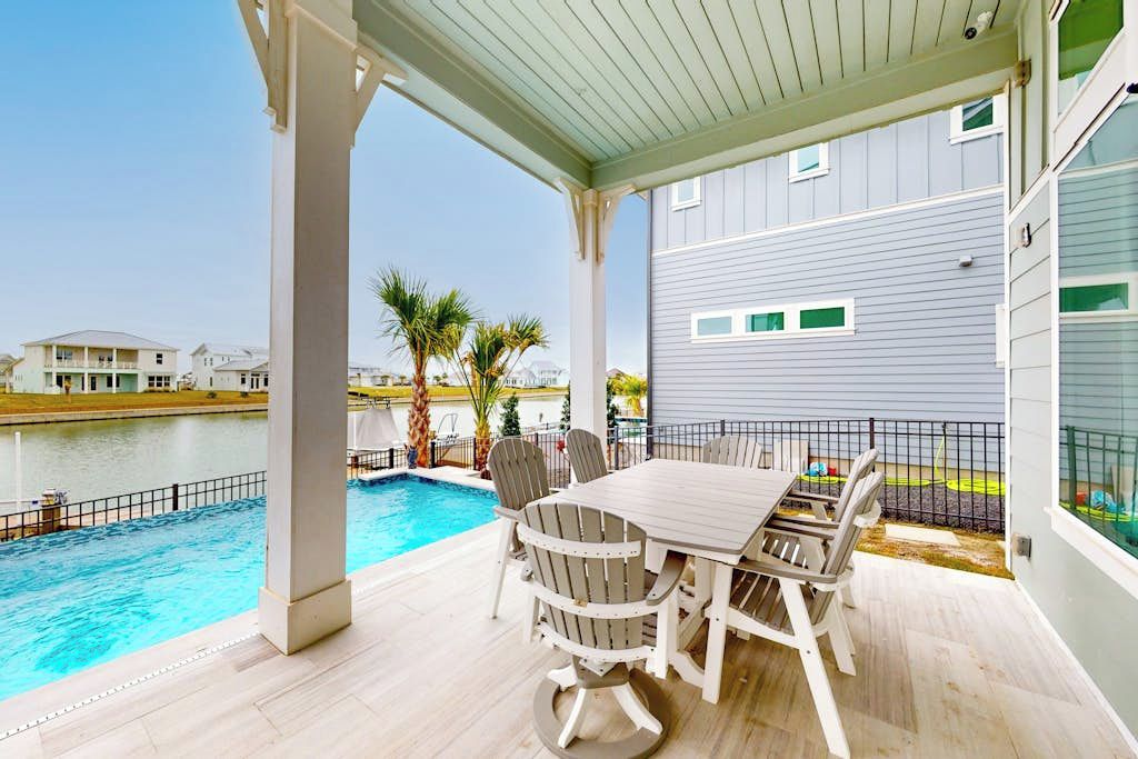 A covered outdoor area with a pool, a table with chairs, and a view of a body of water and buildings.