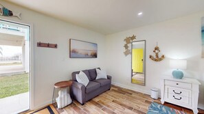 A living room with a grey sofa, a small round table, a white cabinet, and a large mirror. - Bella Vista #25 - Water Views, Pool & Hot Tub (Port Aransas)