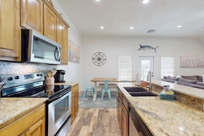 A kitchen with wooden cabinets, a microwave, and a stove. There is a dining area with a table and chairs. - Bella Vista #25 - Water Views, Pool & Hot Tub (Port Aransas)