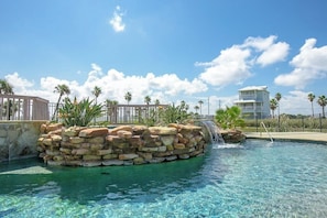 A clear pool with a waterfall feature, surrounded by palm trees and a stone structure. - Bella Vista #25 - Water Views, Pool & Hot Tub (Port Aransas)