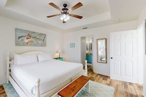 A bedroom with a bed, a ceiling fan, a painting, a door, and a hallway. - Bella Vista #25 - Water Views, Pool & Hot Tub (Port Aransas)