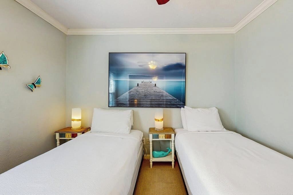 Habitación de hotel con dos camas, una mesita de noche con lámpara, un cuadro colgado en la pared y un pequeño adorno azul turquesa en el suelo.