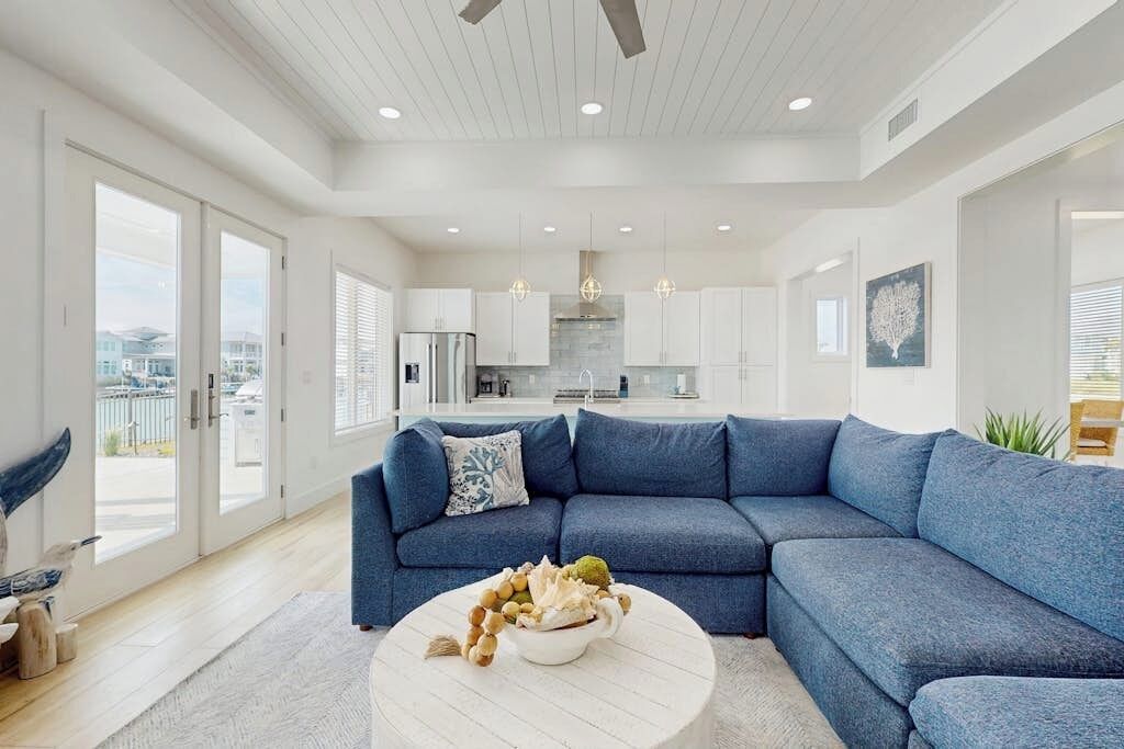 A modern living room with a sectional sofa, a round coffee table with decorative items, and a view of the kitchen and cityscape through large windows.