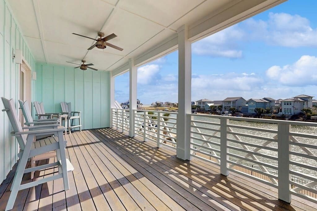 A covered deck with chairs and a railing overlooking a body of water and houses.
