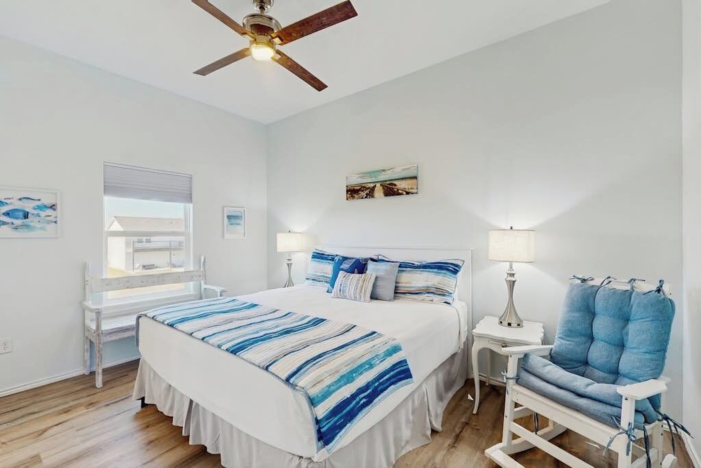 A bedroom with a bed, bedside tables, a chair, a ceiling fan, and artwork on the walls.