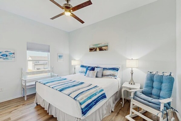 A bedroom with a bed, bedside tables, a chair, a ceiling fan, and artwork on the walls. - Paradise on the Coast (Rockport)