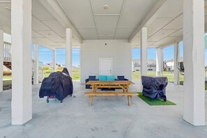 A covered outdoor area with a wooden table and benches, a covered grill, and a door in the background. - Paradise on the Coast (Rockport)