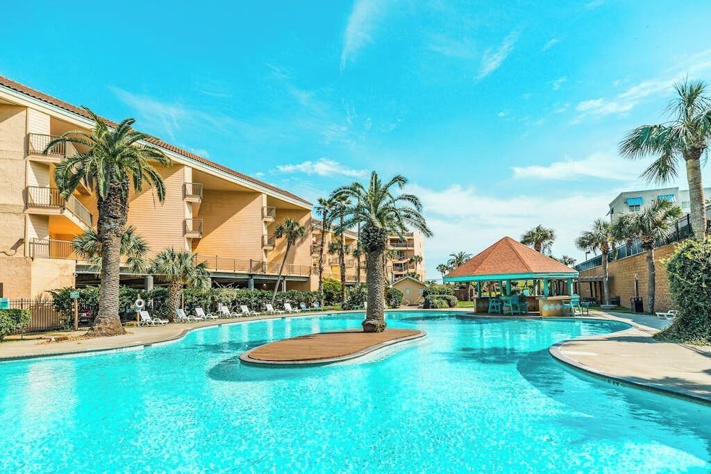 Une piscine entourée de bâtiments d’hôtel, avec des chaises longues et des palmiers.