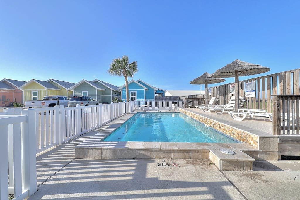 A swimming pool with a white fence, lounge chairs, and umbrellas.
