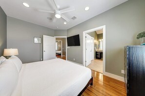 A bedroom with a bed, a ceiling fan, a TV, and a bathroom visible through an open door. - Serenity Island - Two-Story, Pool, Near the Beach (Corpus Christi)