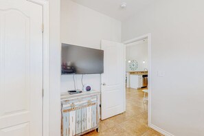 A hallway with a television mounted on the wall, a white door, and a kitchen area visible through an open door. - Morgan Shores - Pool & Garage, Near the Beach (Port Aransas)