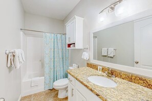 Ein Badezimmer mit einer weißen Toilette, einem Waschbecken mit einer Granitplatte, einer Duschvorhang und einem Spiegel.