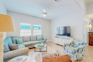 A living room with a sofa, a coffee table, a patterned armchair, a television, and a ceiling fan. - Morgan Shores - Pool & Garage, Near the Beach (Port Aransas)