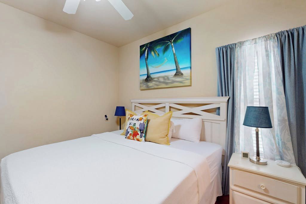 Habitación de hotel con cama, mesita de noche, lámpara, un cuadro de una playa y una ventana con cortinas.