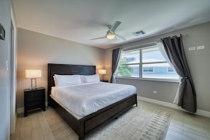 A bedroom with a bed, nightstands, a ceiling fan, a window with curtains, and a view of buildings outside. - Stunning Sunsets & Snorkelling on 7 Mile (George Town)