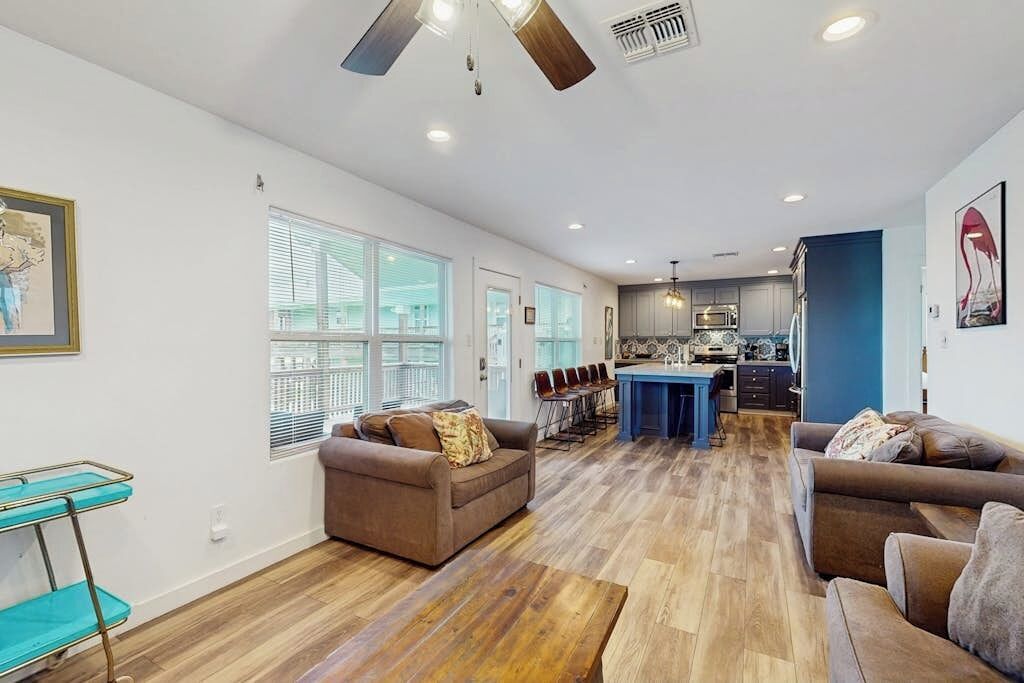 A spacious living area with a kitchen in the background, wooden flooring, and a ceiling fan.