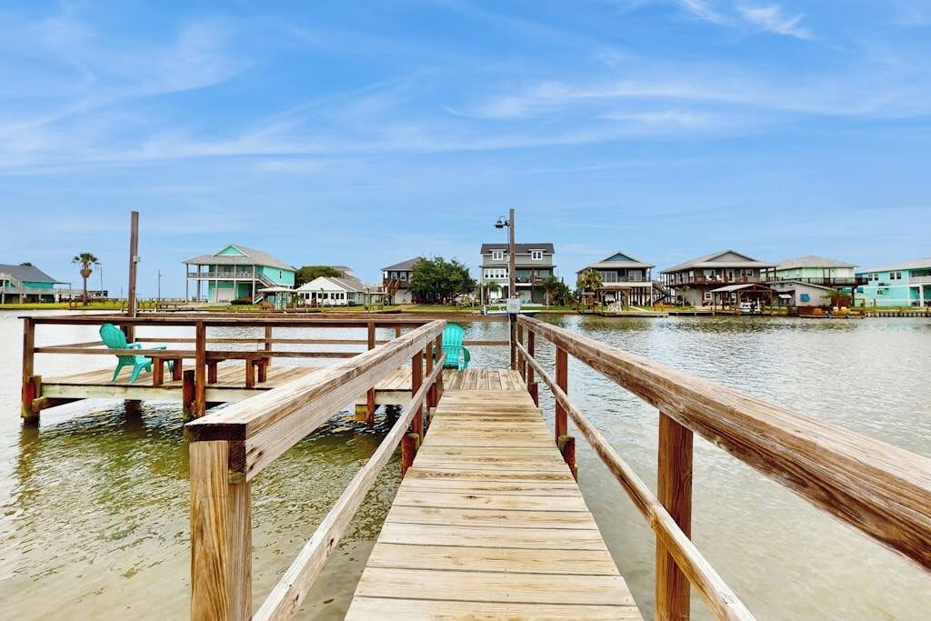A wooden pier leading to a dock with chairs, surrounded by houses with a clear sky above.