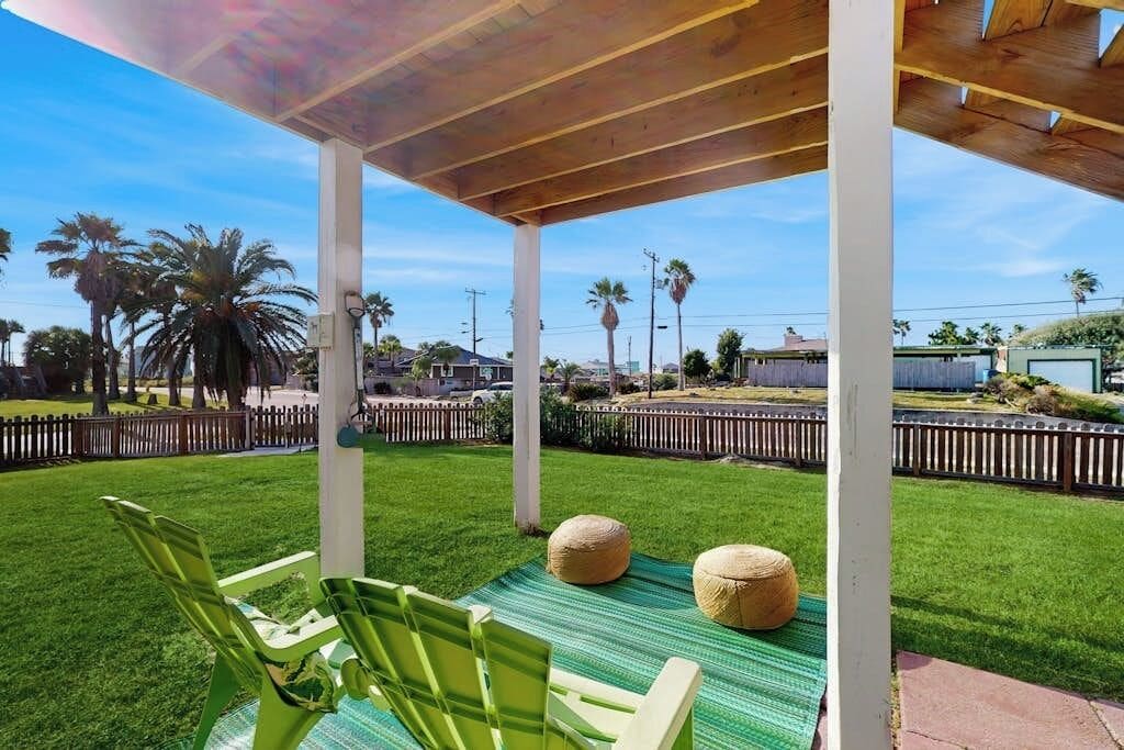 An outdoor patio area with green chairs and a woven ottoman, surrounded by a wooden fence and palm trees.