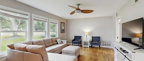 Una sala de estar con un sofá seccional color beige, un ventilador de techo, un televisor y vistas al exterior.