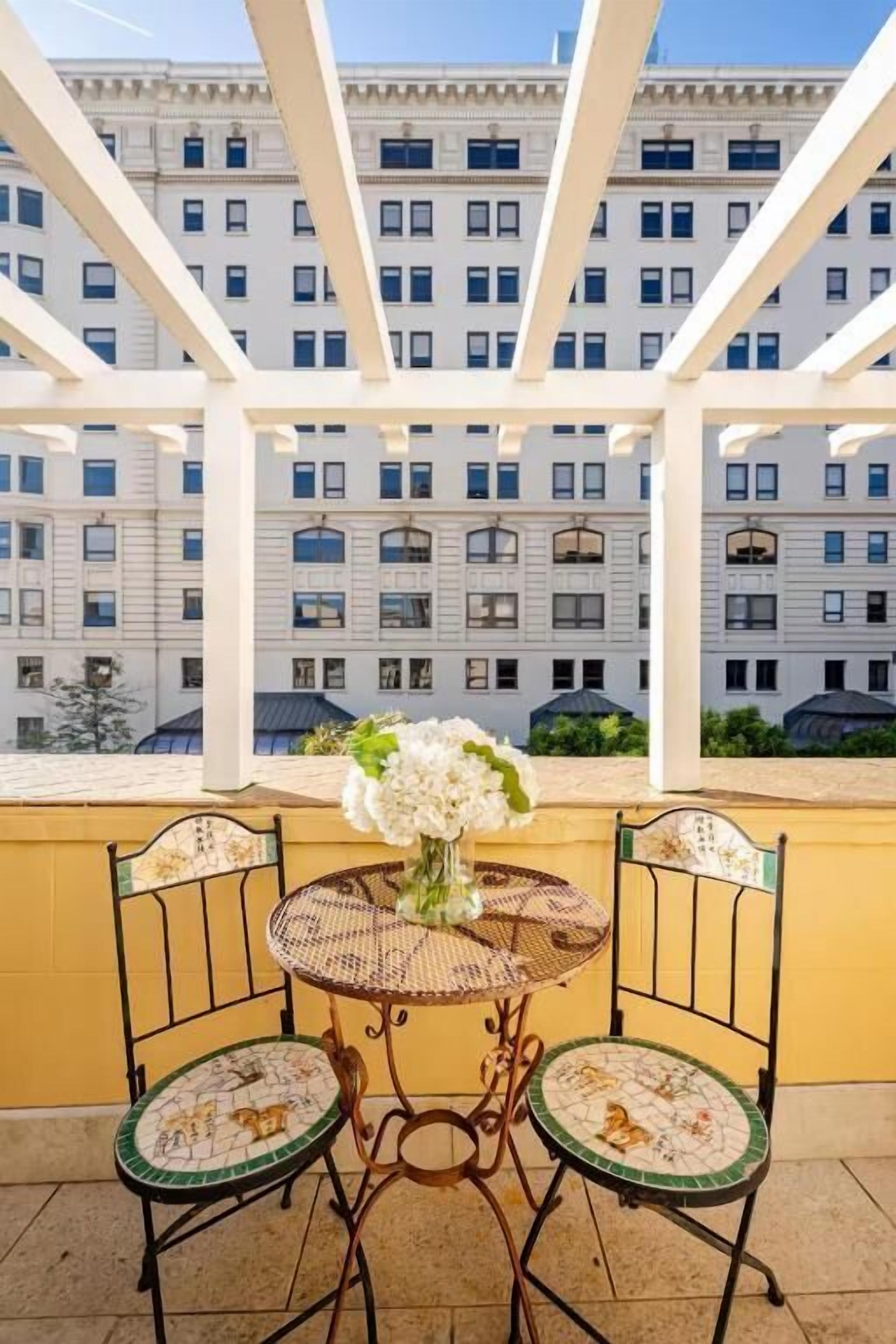 A balcony with a table and chairs, a vase with flowers, and a view of a multi-story building.