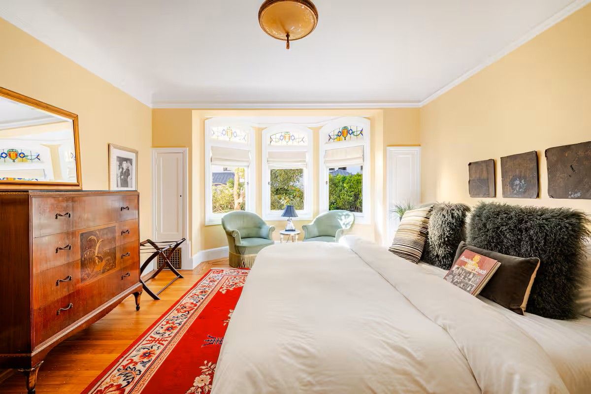 A bedroom with a large bed, a wooden dresser, a red and white patterned rug, and a view of the outdoors through a window.