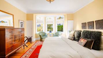 A bedroom with a large bed, a wooden dresser, a red and white patterned rug, and a view of the outdoors through a window.