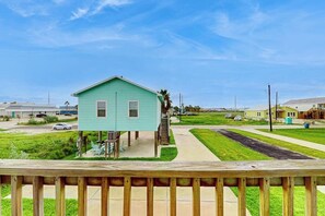 A single-story building with a porch and outdoor seating, surrounded by a grassy area and a clear sky. - Eskridge Gem - Both Homes (Port Aransas)