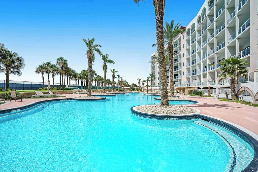 A large outdoor swimming pool with multiple lanes, surrounded by palm trees and a hotel building.