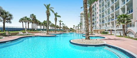 A large outdoor swimming pool with multiple lanes, surrounded by palm trees and a hotel building.