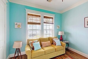 A room with a yellow sofa, two windows with blinds, a lamp, and framed artwork on the walls. - The Conch Casa - Steps to the Beach, Sleeps 9 (Galveston)