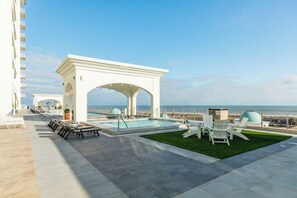 A rooftop pool area with a white arch structure, lounge chairs, and a view of the ocean. - Emerald By The Sea #603 (Galveston)