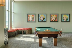 A pool table in a room with a bench, a wall with framed artwork, and a window providing natural light. - Emerald By The Sea #603 (Galveston)
