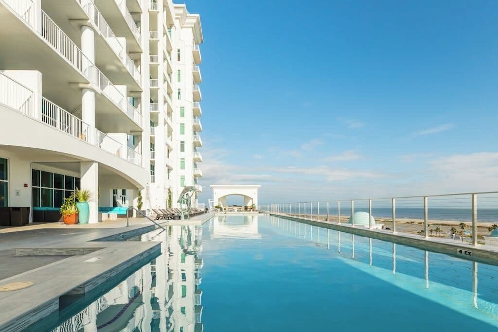 A hotel pool with a clear view of the ocean, a white building with balconies, and a covered walkway.