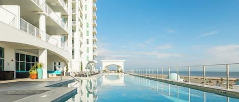 A hotel pool with a clear view of the ocean, a white building with balconies, and a covered walkway.
