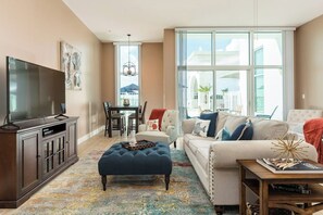 A living room with a television, sofa, ottoman, and a dining area with chairs and a table. - Emerald By The Sea #603 (Galveston)