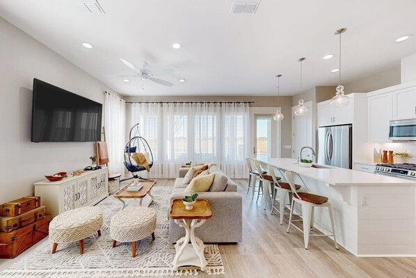Una sala moderna con un sofá, otomanas y una mesa de centro. Hay una zona de cocina con un refrigerador y una mesa de comedor.