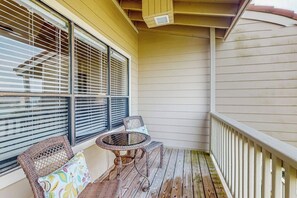 A covered outdoor area with a wooden deck, two wicker chairs, a small round table, and a window with blinds. - Seagull Cove - Waterfront, Bay Views & Garage (Galveston)
