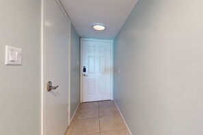 Ein enger Flur mit einer weißen Tür am Ende, einem Lichtschalter an der linken Wand und einer Deckenleuchte.